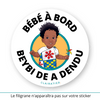 Ce sticker rond illustre un petit garçon noir avec des cheveux bouclés et volumineux (cheveux afro garçon). Il porte un t-shirt blanc avec une grande fleur rouge stylisée sur le devant, accompagné d’un pagne coloré en bleu, jaune et rouge noué sur son épaule gauche. Dans sa main droite, il tient un moulin à vent multicolore.  Le texte "Beybi de a dendu", en langue bushinengué (Saramaka -  langue notamment parlée au Suriname et Guyane française), est inscrit en bas en noir