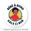 Ce sticker rond représente un petit garçon noir avec des cheveux bouclés et volumineux (cheveux afro garçon). Il porte un t-shirt rouge uni et tient un moulin à vent multicolore dans sa main droite. Le texte "Bébé à Bord" est inscrit en haut, tandis que "Xale Ci Biir", en wolof (langue notamment parlée au Sénégal et en Mauritanie), apparaît en bas en noir. Le texte "Clairatier" est situé au bas du sticker. Sticker bébé à bord.