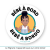 Ce sticker rond illustre une petite fille noire avec des cheveux coiffés en un chignon haut volumineux et une frange bouclée sur le devant. Elle porte une robe blanche à rayures bleues ornée d’un petit cœur rouge sur le devant. Dans sa main droite, elle tient un moulin à vent multicolore. Le texte "Bébé à Bord" est inscrit en haut, tandis que "Bebê a Bordo", en portugais (langue parlée dans des pays comme le Brésil, le Portugal ou encore le Cap-Vert), apparaît en bas en noir.  Sticker bebê a bordo.